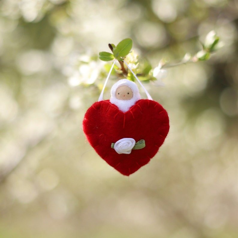 May include: A handmade felt heart ornament in vibrant red, featuring a small doll with a white hood and a delicate white rose accent. The ornament hangs from a white ribbon, perfect for Valentine's Day or a special occasion.