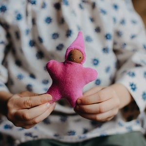 May include: A child holds a pink felt star-shaped doll with a brown face and a pink pointed hat.