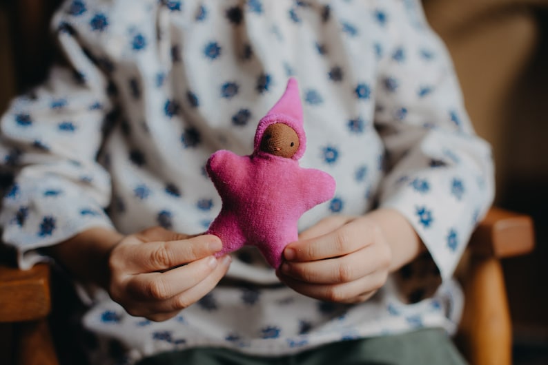 May include: A child holds a pink felt star-shaped doll with a brown face and a pink pointed hat.