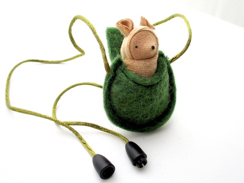 May include: A green felt pouch with a drawstring closure, containing a small stuffed animal with a beige body and a brown nose. The pouch is attached to a green cord with black cord ends.