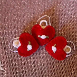 May include: Three red felt heart ornaments with white ribbon handles. Each heart has a small white rose and green ribbon detail. The ornaments are arranged on a textured, patterned surface.