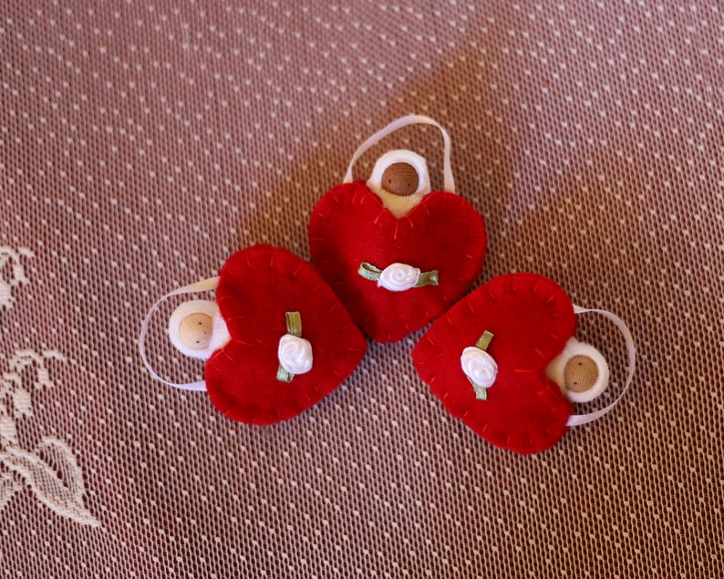 May include: Three red felt heart ornaments with white ribbon handles. Each heart has a small white rose and green ribbon detail. The ornaments are arranged on a textured, patterned surface.