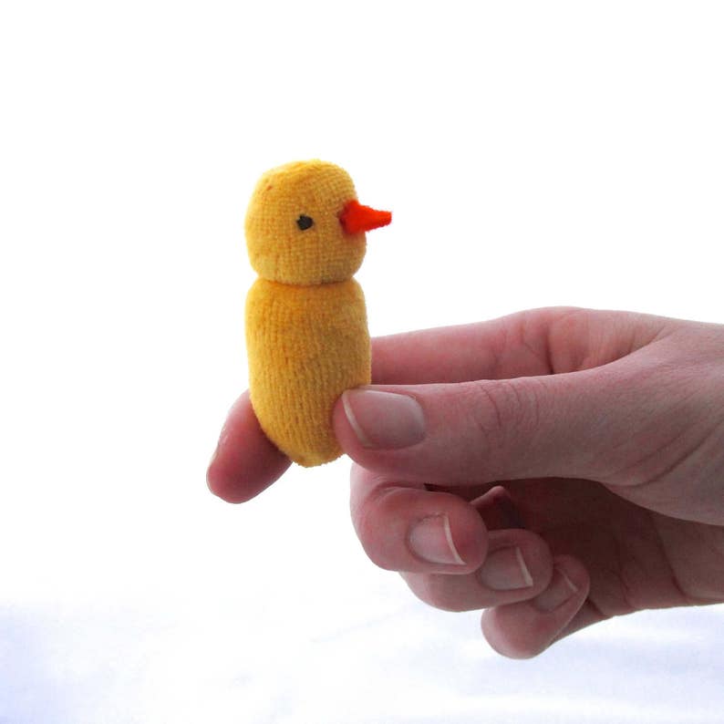 Op de afbeelding: Een kleine, handgemaakte gele kuiken knuffel met een oranje snavel en een zwart oog. Het kuiken is gemaakt van een zacht, getextureerd materiaal en wordt in de hand van een persoon tegen een witte achtergrond gehouden. Het speelgoed is ongeveer 5 cm hoog.