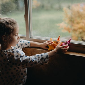 May include: A young girl plays with colorful felt finger puppets near a window. The puppets are shaped like people with pointy hats.