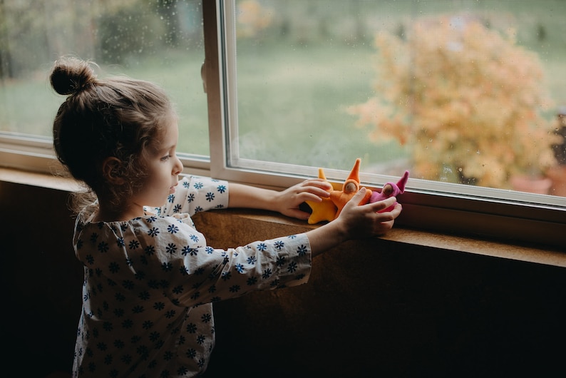 May include: A young girl plays with colorful felt finger puppets near a window. The puppets are shaped like people with pointy hats.
