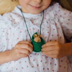 May include: A young girl wearing a white dress with a red floral pattern holds a green felt pouch with a small stuffed animal inside. The pouch is attached to a green cord and worn as a necklace.