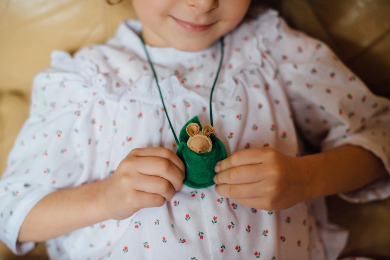 May include: A young girl wearing a white dress with a red floral pattern holds a green felt pouch with a small stuffed animal inside. The pouch is attached to a green cord and worn as a necklace.