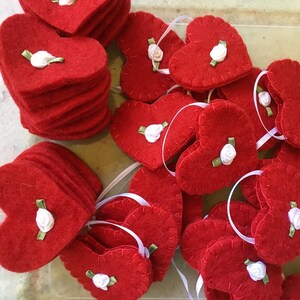 May include: Red felt heart ornaments with white ribbon hangers. Each heart is decorated with a small white rose and green ribbon. Some hearts are stacked, while others are scattered.