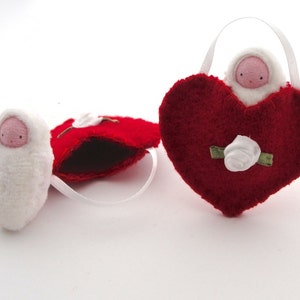 May include: Three handmade felt ornaments. One is a white swaddled baby doll. Another is a red heart-shaped ornament with a white rose and a baby doll inside. The third is a red heart-shaped ornament with a white rose.