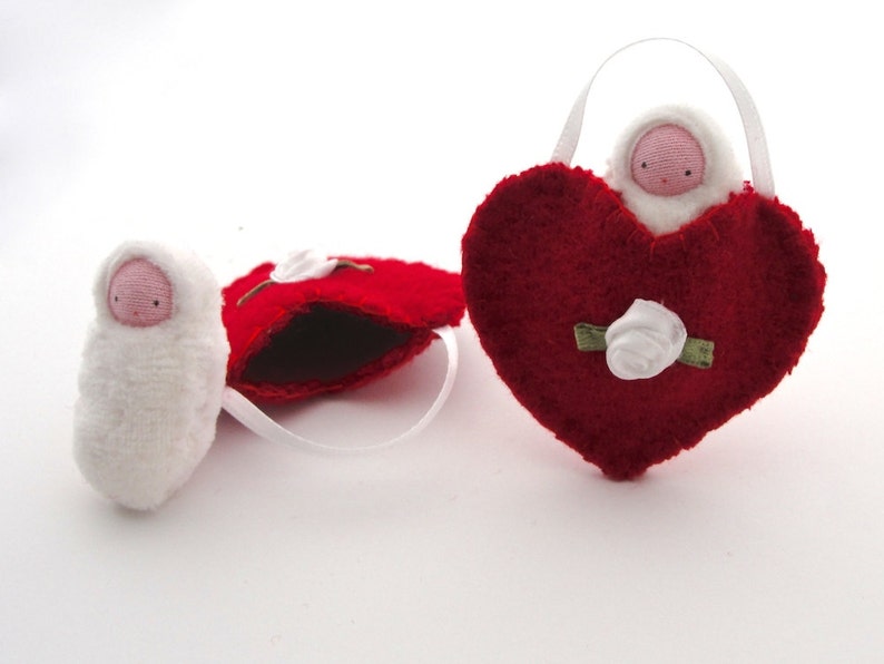 May include: Three handmade felt ornaments. One is a white swaddled baby doll. Another is a red heart-shaped ornament with a white rose and a baby doll inside. The third is a red heart-shaped ornament with a white rose.