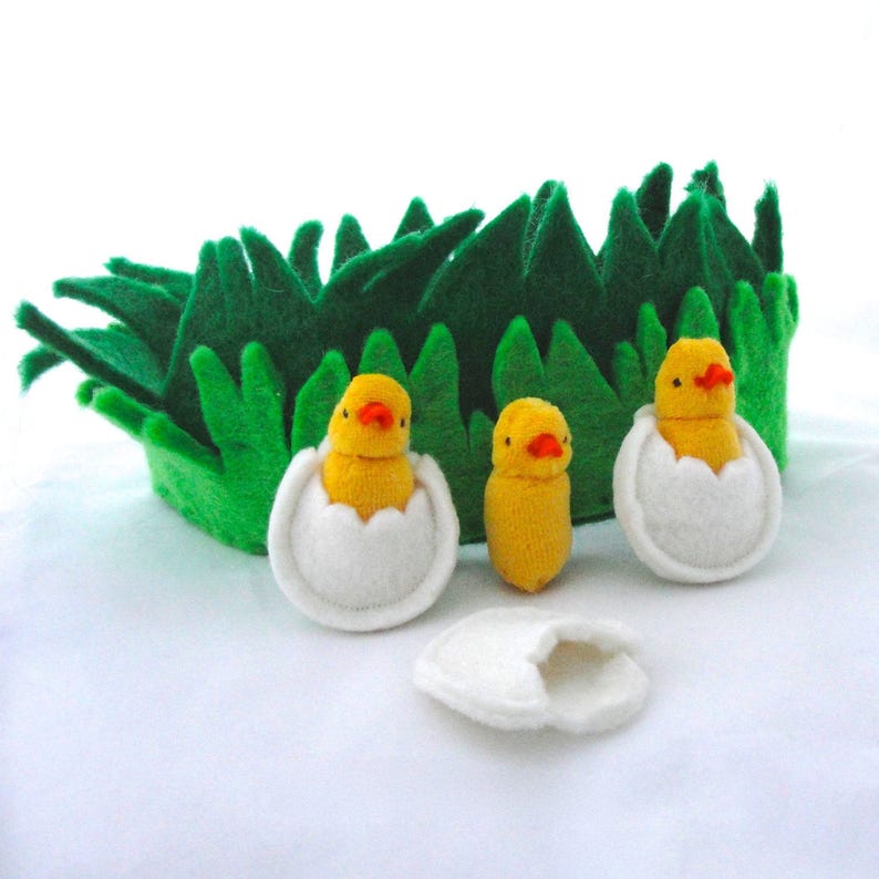 Op de afbeelding: Een vilten Paassc&egrave;ne met een groene grasachtergrond, drie gele kuikens en twee witte eierhelften. E&eacute;n kuiken staat, twee zitten in eierschalen. De kuikens hebben oranje snavels.