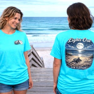 May include: Two women wearing turquoise t-shirts with turtle designs. One shirt has a small turtle graphic, the other has a full moon and turtle scene with the text "Lights Out for Turtles". The background is a beach.