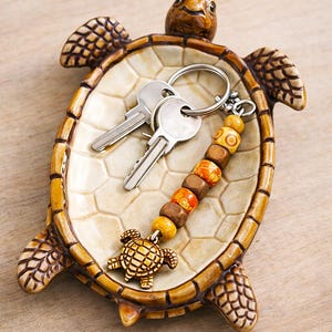 以下が含まれることがあります： クリーム色の内側と茶色の縁取りが施された、カメの形をしたセラミック製の小物入れ。2つの銀色の鍵と、カメのチャームが付いたビーズのキーチェーンが入っています。キーチェーンは、茶色、オレンジ色、黄色の木製ビーズが特徴です。