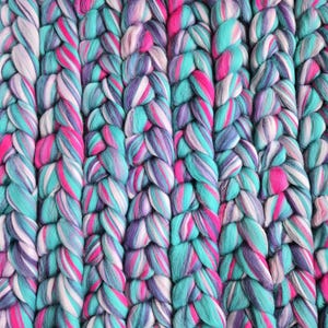 May include: Close-up of a chunky knit textile in a vertical braid pattern. The yarn is a mix of colours, including teal, pink, purple, and white. The texture appears soft and inviting, suitable for a blanket or throw.
