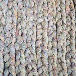 Könnte beinhalten: Nahaufnahme eines grob gestrickten Textils in einer neutralen Farbe mit blauen, rosa, gelben und grünen Tupfen. Das dicke Garn ist in einem geflochtenen Muster gewebt und erzeugt eine strukturierte Oberfläche. Der Gesamteffekt ist weich und einladend.