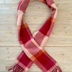 May include: A long rectangular scarf featuring a plaid pattern in shades of deep red, bright pink, soft light pink, and orange. The scarf is finished with fringed ends consisting of loose threads in matching red and light pink color tones.