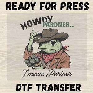 May include: DTF transfer with a cartoon frog in cowboy attire: brown hat, jacket, and red bandana. The frog holds a cactus pot. Text includes "READY FOR PRESS", "HOWDY PARDNER...", "I mean, Partner", and "DTF TRANSFER".