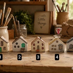 Może przedstawiać: Pięć miniaturowych domków ceramicznych ustawionych w rzędzie, każdy z unikalnymi detalami architektonicznymi, oknami i drzwiami. Dom 1 ma zielone drzwi, dom 2 zielone łukowe drzwi, dom 3 brązowe drzwi, dom 4 różowy balon, a dom 5 szare drzwi.