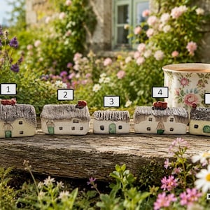 Può includere: Cinque statuette di case in miniatura in ceramica numerate da uno a cinque, ognuna caratterizzata da un tetto di paglia testurizzato, pareti bianco sporco e una piccola porta verde, disposte in fila accanto a una tazza da tè a motivi floreali.