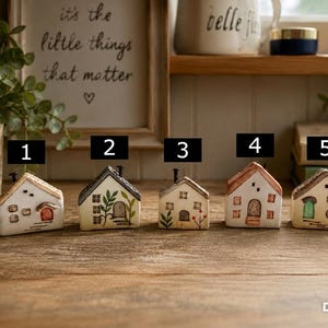 Może przedstawiać: Pięć małych ceramicznych figurek domków ustawionych w rzędzie, każdy ponumerowany od jednego do pięciu. Każdy domek ma unikalny, ręcznie malowany wzór, w tym różne kolory drzwi, kształty okien i motywy botaniczne, zwieńczone małym czarnym kominem.