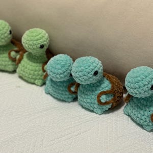 May include: A row of handmade crochet turtle figurines in shades of green and teal. Each turtle has a rounded head, a small body, and brown accents. The turtles are arranged on a white surface.