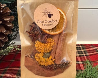May include: A clear cellophane bag filled with chai potpourri. The bag contains dried orange slices, cinnamon sticks, star anise, and yellow flowers. A round label with the text "Chai Comfort Potpourri" is affixed to the bag. The bag is tied with twine.