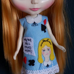 May include: A doll with long, orange hair wearing a light blue dress with a white collar and lace trim. The dress features a drawing of a blonde girl and the text "WE'RE ALL MAD HERE!" along with playing card symbols.