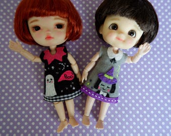 Puede incluir: Dos muñecas, una pelirroja y otra morena, vestidas con trajes de Halloween. Una muñeca tiene un vestido negro con un fantasma y la palabra "Boo". La otra muñeca lleva un vestido gris con un sombrero de bruja.