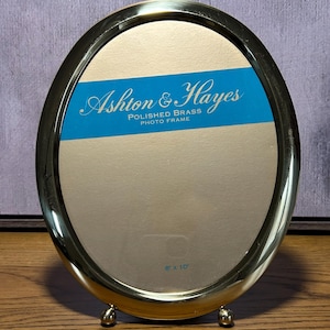 May include: Oval polished brass photo frame with the text "Ashton & Hayes". The frame is supported by small ball feet. The frame is 8 x 10 inches.