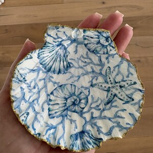 May include: A decorative scallop shell dish featuring a white base with a blue printed pattern of seashells, starfish, and coral branches. The scalloped edges of the shell are finished with a metallic gold trim, creating a distinct border.