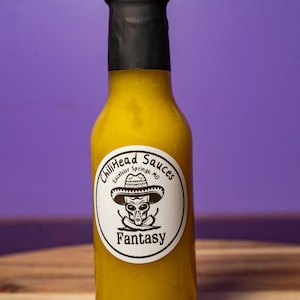 Könnte beinhalten: Eine Flasche gelber scharfer Sauce mit schwarzem Verschluss. Das Etikett trägt die Aufschrift "ChiliHead Sauces Excelsior Springs, MO" mit einer Grafik eines Aliens mit Sombrero. Das Wort "Fantasy" steht darunter.