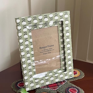 May include: A rectangular picture frame with a floral pattern in white on a light green background. The frame has a clear glass insert and the text "Bangor Frame" is visible. The frame is resting on a table with decorative coasters.