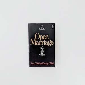 May include: A black paperback book titled Open Marriage: A New Life Style for Couples by Nena O'Neill and George O'Neill. The cover features gold serif lettering, the text The #1 Bestseller, and a small white logo in the top right corner.