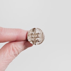 May include: A silver-toned ring with a textured, circular design. The ring features a series of vertical and horizontal lines, creating a geometric pattern. The ring is held against a plain white background.