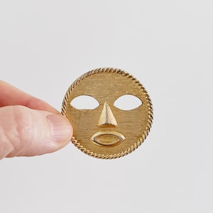 May include: A gold-colored metal mask with a simple face design. The mask has two eye holes, a triangular nose, and a small oval mouth.