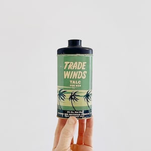 May include: A green and black metal container with a black lid. The container has a label that reads "TRADE WINDS TALC FOR MEN" and features a palm tree design. The container is 3 1/2 ounces.
