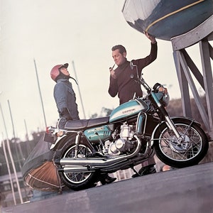 May include: A vintage poster advertising the Suzuki GT750 motorcycle. The poster features a green motorcycle with two men standing next to it. The motorcycle is parked on a ramp with a sailboat in the background.