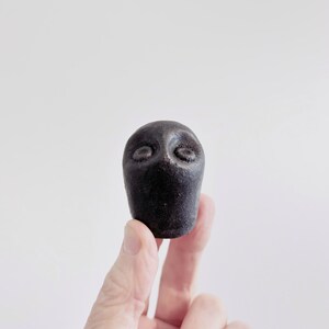 May include: A small, dark, textured object shaped like a stylized head with two circular indentations for eyes. The surface has a matte, granular appearance, and the form is rounded at the top, tapering slightly toward the base.