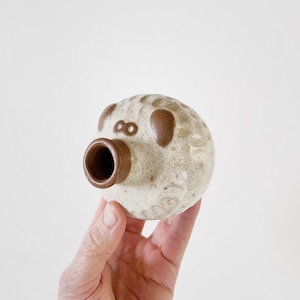 May include: A small, white ceramic figurine shaped like a pig with brown accents. The pig has a round body and a small snout with a hole in the center.