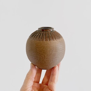 May include: A small, round, brown ceramic vase with a textured surface. The vase has a slightly darker brown band around the top.