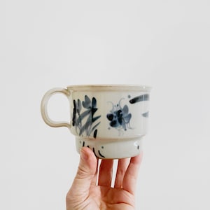 May include: Cream-colored ceramic mug with a handle, featuring hand-painted blue floral designs. The mug has a stacked design with a smaller base. The mug is held by a hand against a white background.