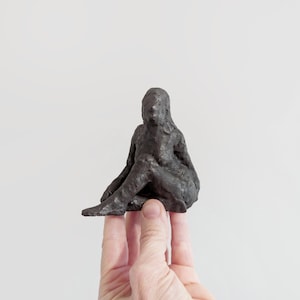 May include: A small, dark gray, textured sculpture of a seated figure with a rough, hand-molded appearance. The figure is depicted in a cross-legged position with a rounded head and defined torso, showcasing a matte finish and uneven surface details.