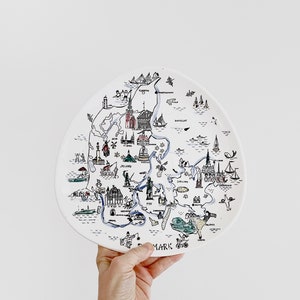 以下が含まれることがあります： デンマークの手描きの地図が描かれた白いセラミックプレート。黒、青、赤でランドマークやイラストが描かれています。「MARK」の文字がプレートに印刷されています。