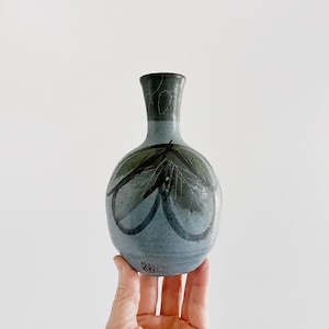 May include: A small, round ceramic vase with a narrow neck, featuring a light blue glaze base. The surface is decorated with dark, hand-painted leaf motifs and abstract black line work. A small, circular maker's mark is visible near the base.