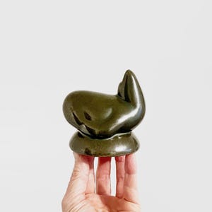 May include: A dark green ceramic sculpture with an abstract design. The artwork features a rounded base and a curved, organic form. The sculpture is held in a hand against a white backdrop.