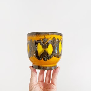 May include: A ceramic plant pot with a striking design. The pot has a base and a broad, rounded body. The exterior is decorated with a pattern of yellow and brown shapes against an orange backdrop. The pot is held by a hand.