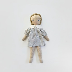 May include: A fabric doll with light tan skin, brown yarn hair, and blue embroidered eyes. The doll wears a white collar and a short-sleeved dress featuring a small blue and white floral pattern. The arms and legs are simple, unjointed fabric tubes.