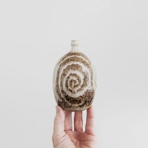 May include: A small, oval-shaped ceramic vase with a narrow neck and a textured surface. The piece features a prominent brown and cream-coloured spiral pattern that wraps around the body, with a mottled, earthy finish near the base.