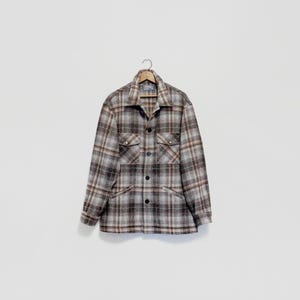 May include: A long-sleeve button-up shirt featuring a plaid pattern in shades of grey, brown, and white. The shirt includes a pointed collar, two chest pockets with buttoned flaps, and a full-length front button closure on a wooden hanger.