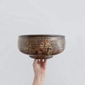 May include: A round ceramic bowl featuring a mottled brown and tan glaze with a textured, earthy finish. The bowl has a wide, open top and a slightly tapered base, showcasing a rustic appearance with varied dark and light brown color tones.